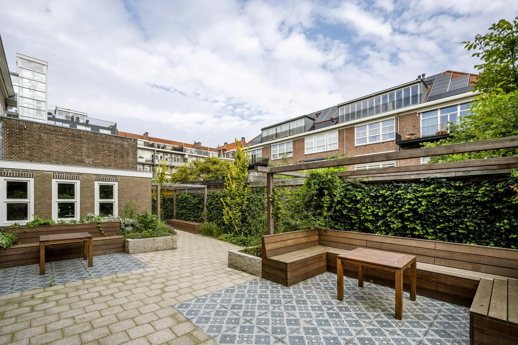 Gemeenschappelijke binnentuin aan de Hillegomstraat met houten zitbanken, tafels, betegeld terras en groene begroeiing.