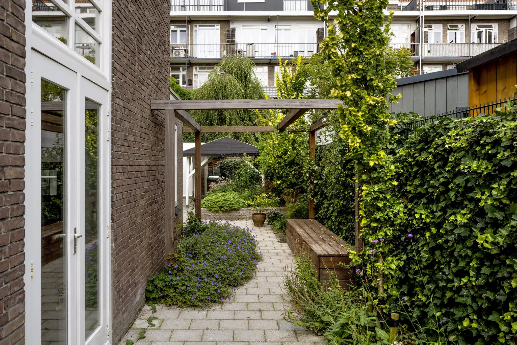 Sfeervolle, smalle stadstuin aan de Hillegomstraat met pergola, groene beplanting en houten zitbank.