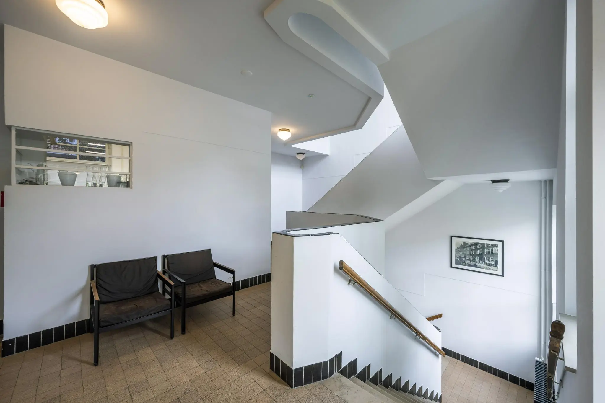 Trappenhuis met strakke witte muren, zwart-witte tegelrand, twee stoelen en een foto aan de muur in een gebouw aan de Hillegomstraat.