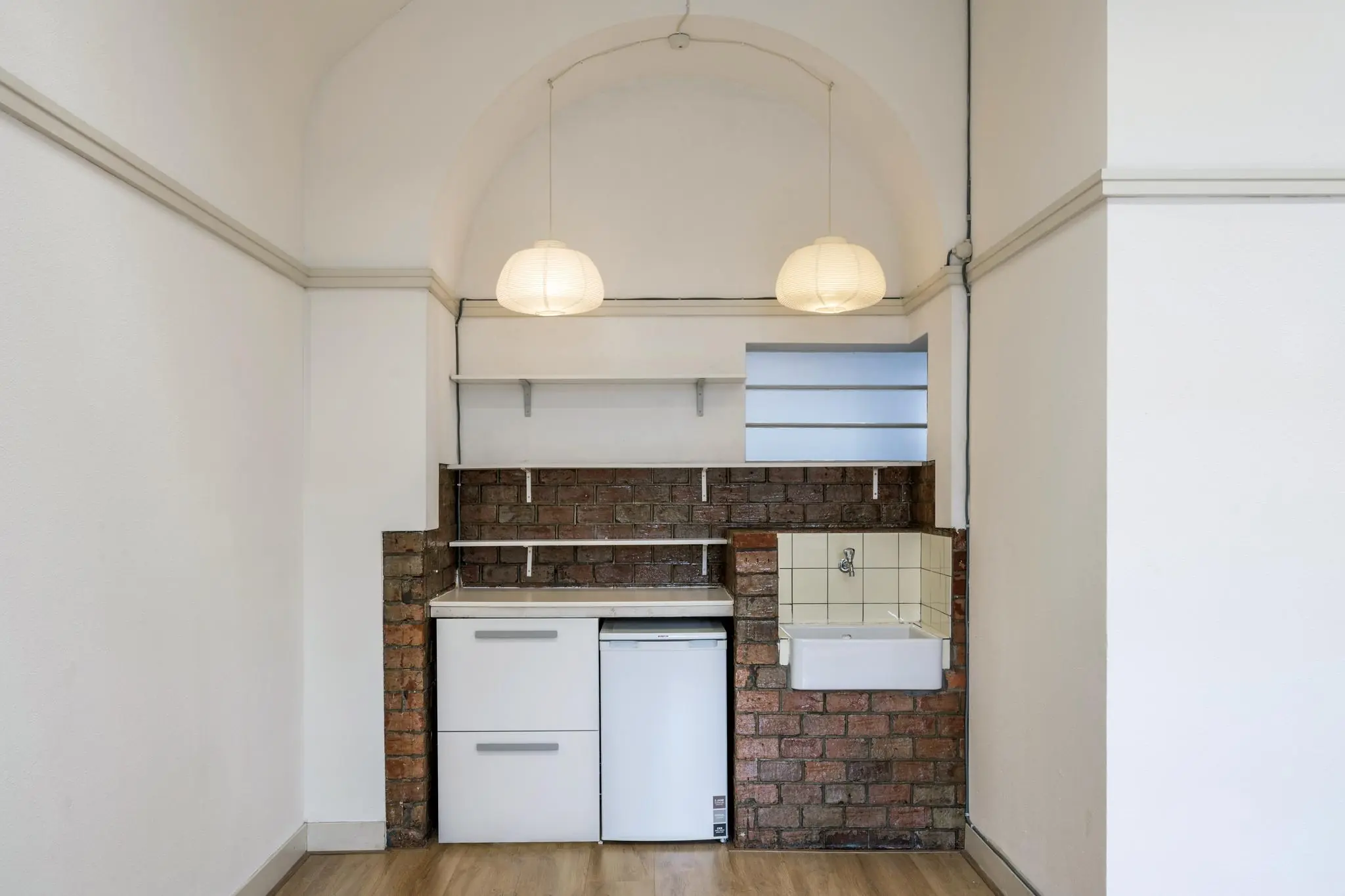 Kleine kitchenette met witte kastjes, bakstenen achterwand, spoelbak en twee hanglampen in een nis aan de Hillegomstraat.
