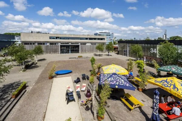 Zonnig plein aan de Johan van Hasseltweg met kleurrijke parasols, picknicktafels en een modern industrieel gebouw op de achtergrond.