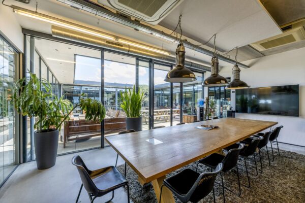 Vergaderruimte aan de Johan van Hasseltweg met houten tafel, moderne verlichting, grote planten en uitzicht op een zonnige binnenplaats.