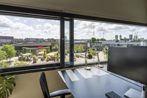 Uitzicht vanuit een modern kantoorraam aan de Johan van Hasseltweg op een zonnig plein met terras en het gebouw PARK.