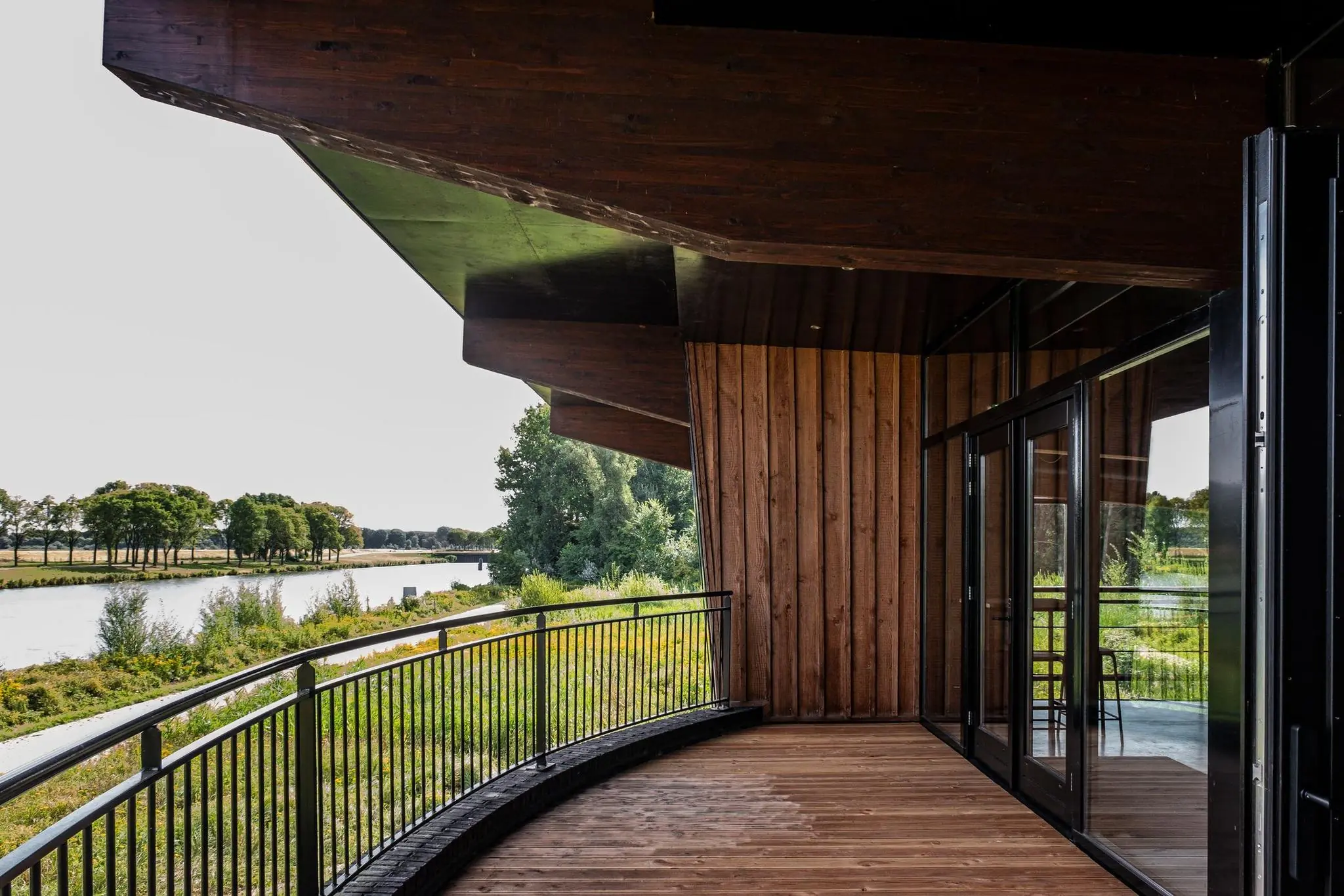 Houten terras met glazen schuifdeuren en uitzicht op een rivier en groene oever langs de Plaatweg.