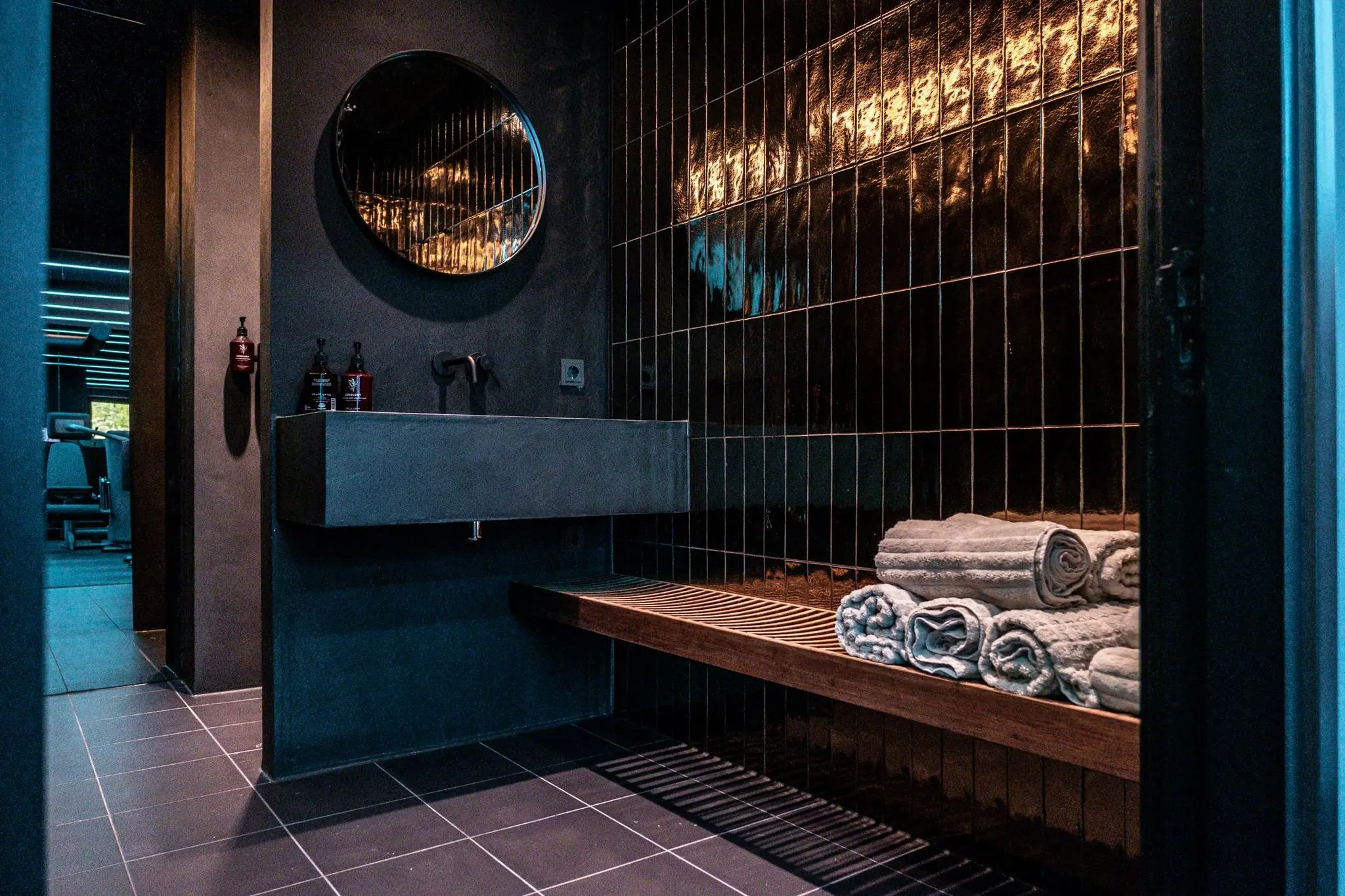 Moderne, donkere wasruimte met een betonnen wastafel, ronde spiegel, stapel opgerolde handdoeken en glanzende zwarte wandtegels.