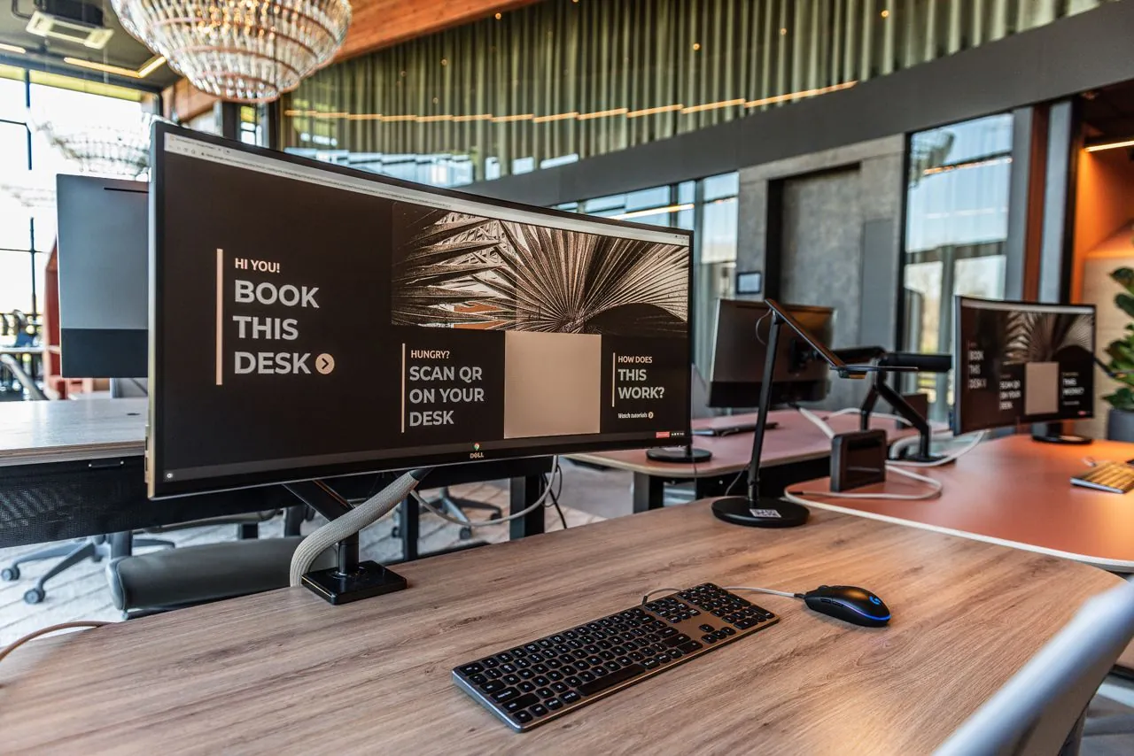 Moderne flexwerkplek met beeldscherm waarop staat "Book this desk" in een open en stijlvol kantoor.