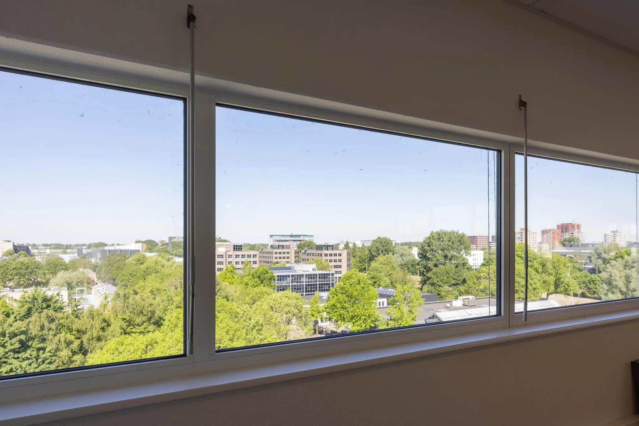 Uitzicht vanuit een raam op de Overschiestraat met veel groen en kantoorpanden in de verte.