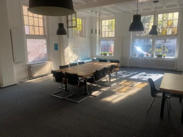 Vergaderruimte aan de Gasthuisplaats met grote ramen, houten tafel, zwarte stoelen en invallend zonlicht.