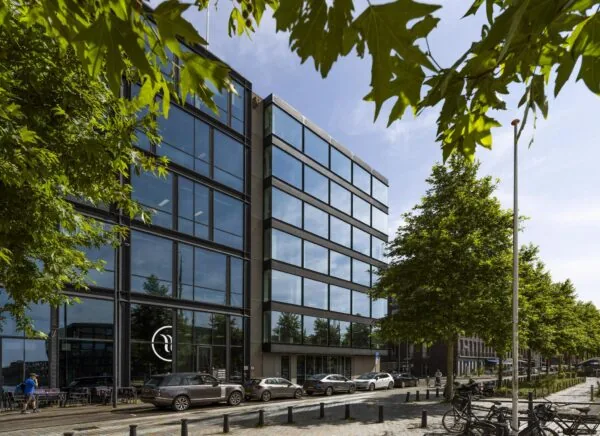 Modern kantoorgebouw aan de Krijn Taconiskade met glazen gevel en geparkeerde auto's langs een boomrijke straat.