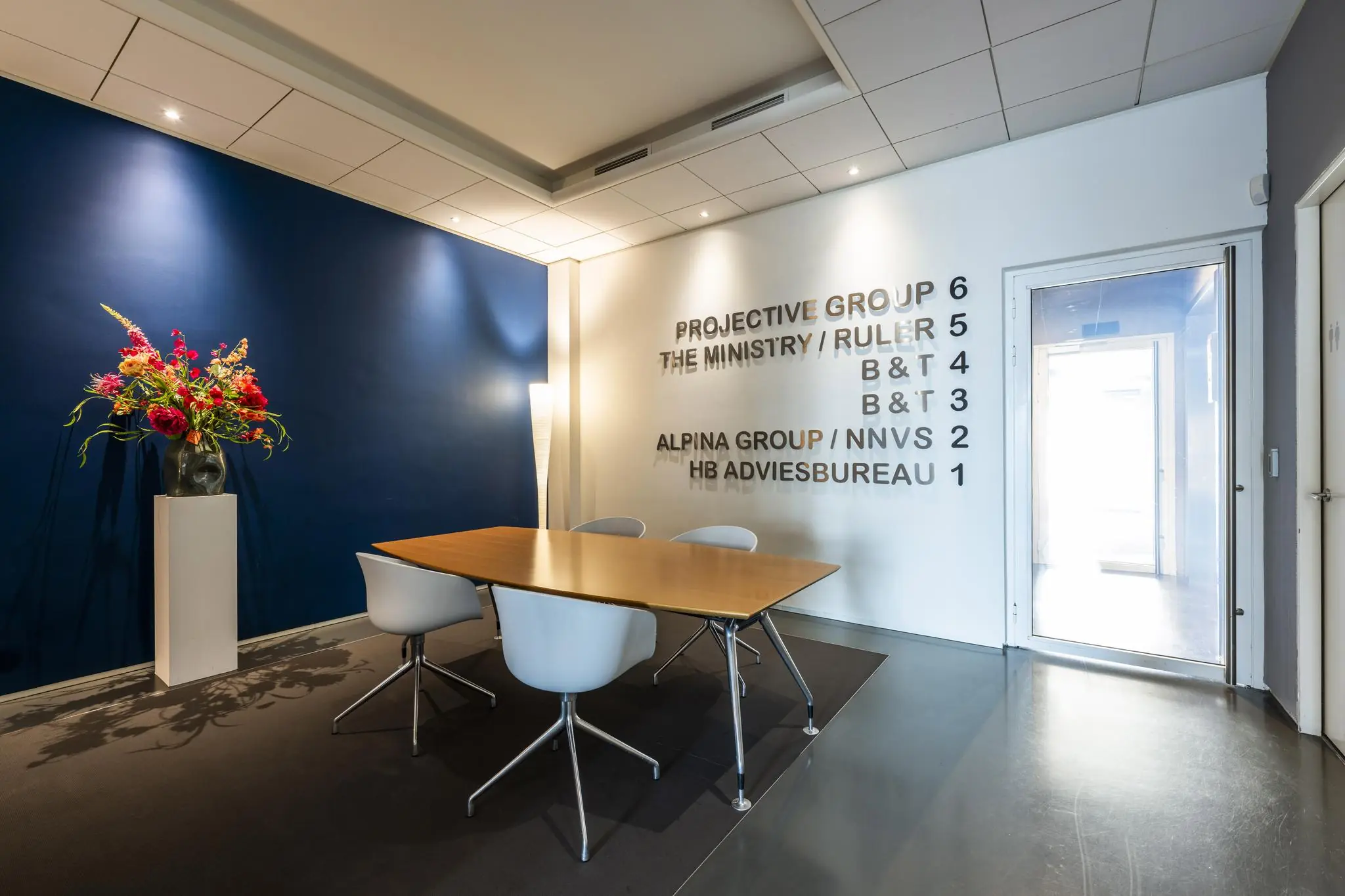 Moderne ontvangstruimte aan de Krijn Taconiskade met houten vergadertafel, stoelen, een donkerblauwe wand met bloemenvaas en namen van bedrijven op de witte muur.