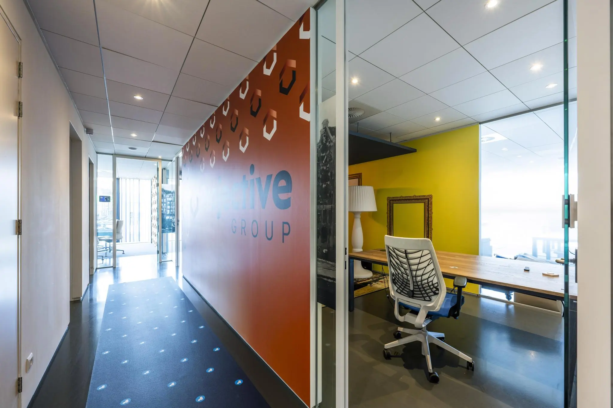 Moderne kantoorruimte aan de Krijn Taconiskade met een glazen vergaderruimte, felgele muur en opvallend oranje wand met het logo van Objective Group.