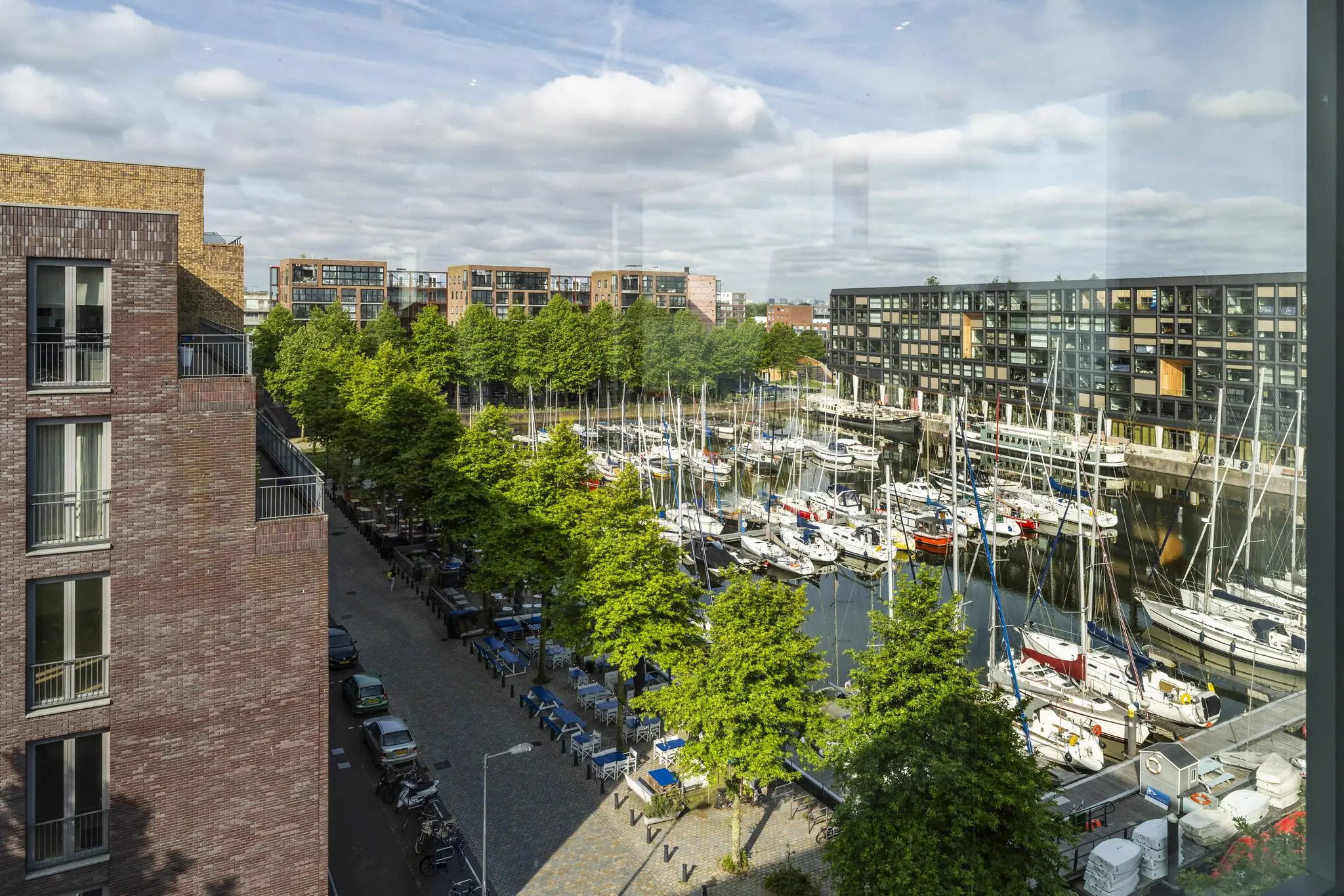 Uitzicht op de jachthaven aan de Krijn Taconiskade met aangemeerde zeilboten, omringd door moderne woongebouwen en groene bomen.