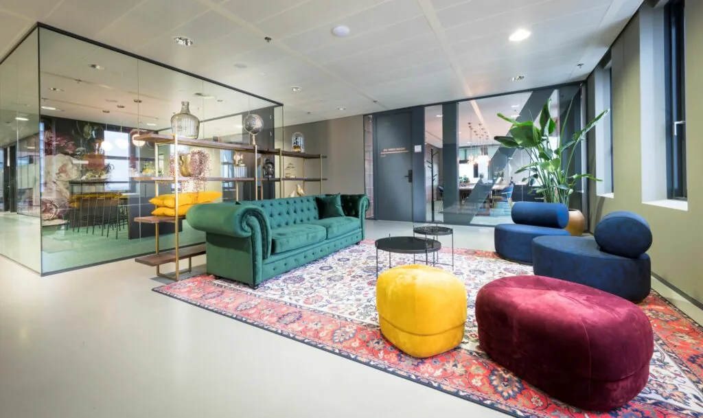 Stijlvolle en kleurrijke loungehoek in een modern kantoor met een groene bank, designmeubels en een glazen vergaderruimte op de achtergrond.