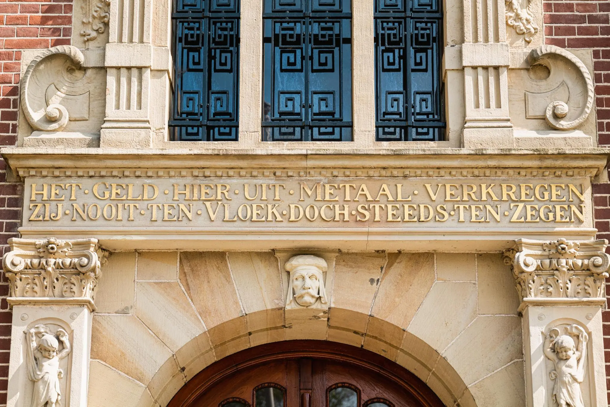 Geveldetail van het pand aan de Leidseweg met een inscriptie over metaal en zegen boven een monumentale houten deur.
