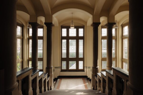 Trapzaal met glas-in-loodramen en zuilen in een historisch gebouw aan de Leidseweg.