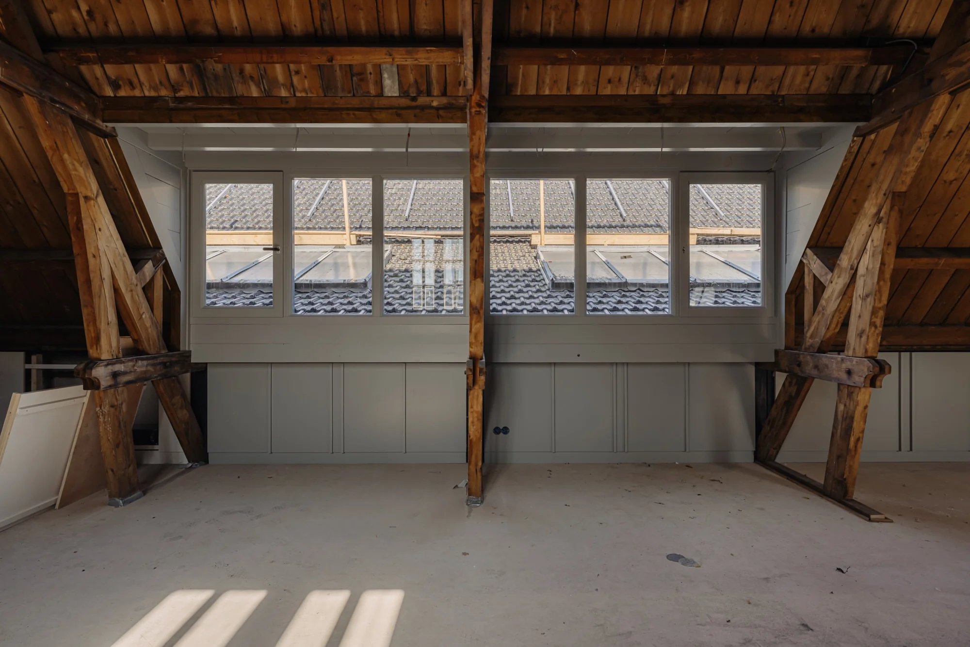 Zolderruimte met houten balken en een rij witte ramen met uitzicht op de dakpannen van Leidseweg 175.