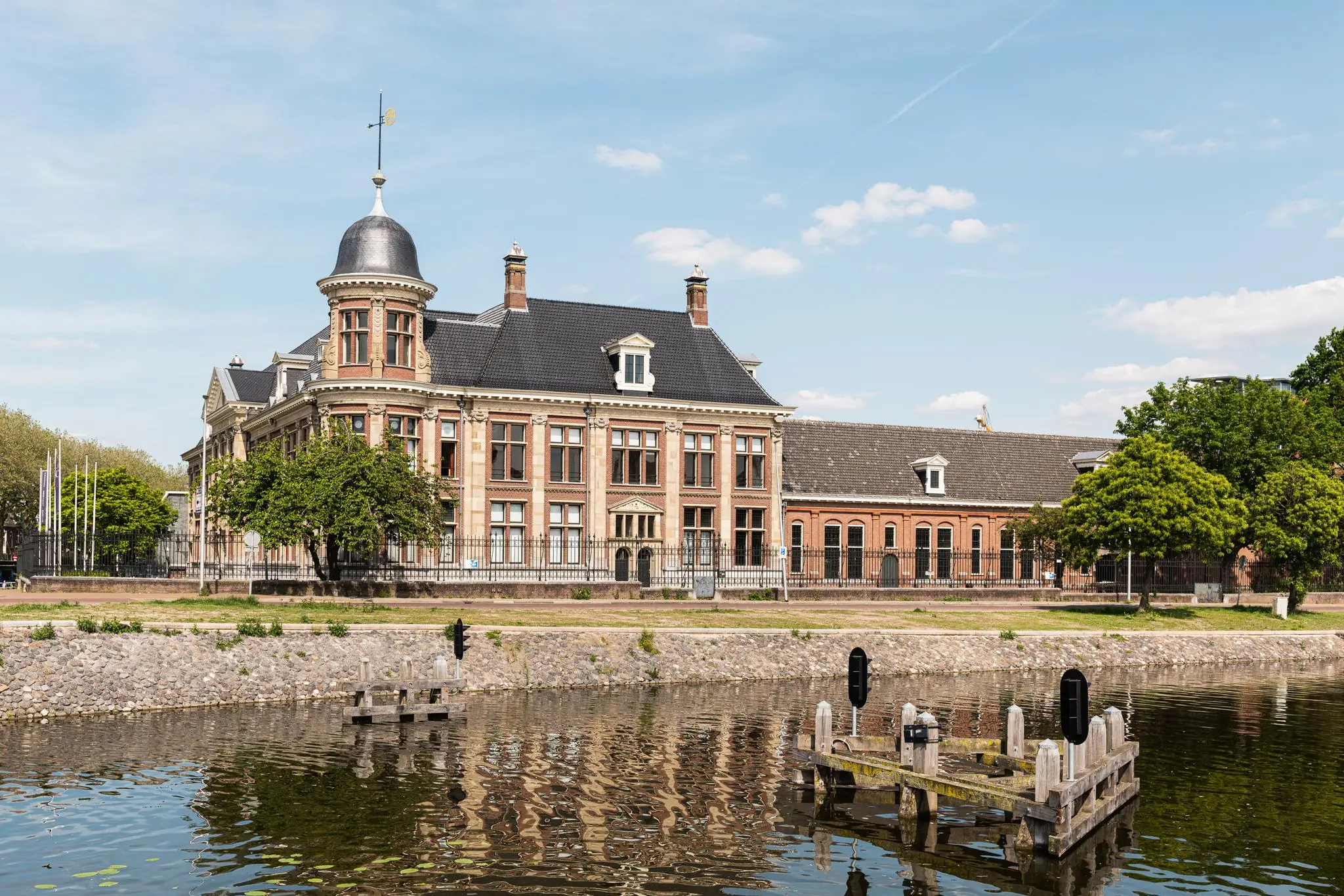 Monumentaal pand aan de Muntkade in Utrecht, met torentje en weerspiegeling in het water op een zonnige dag.