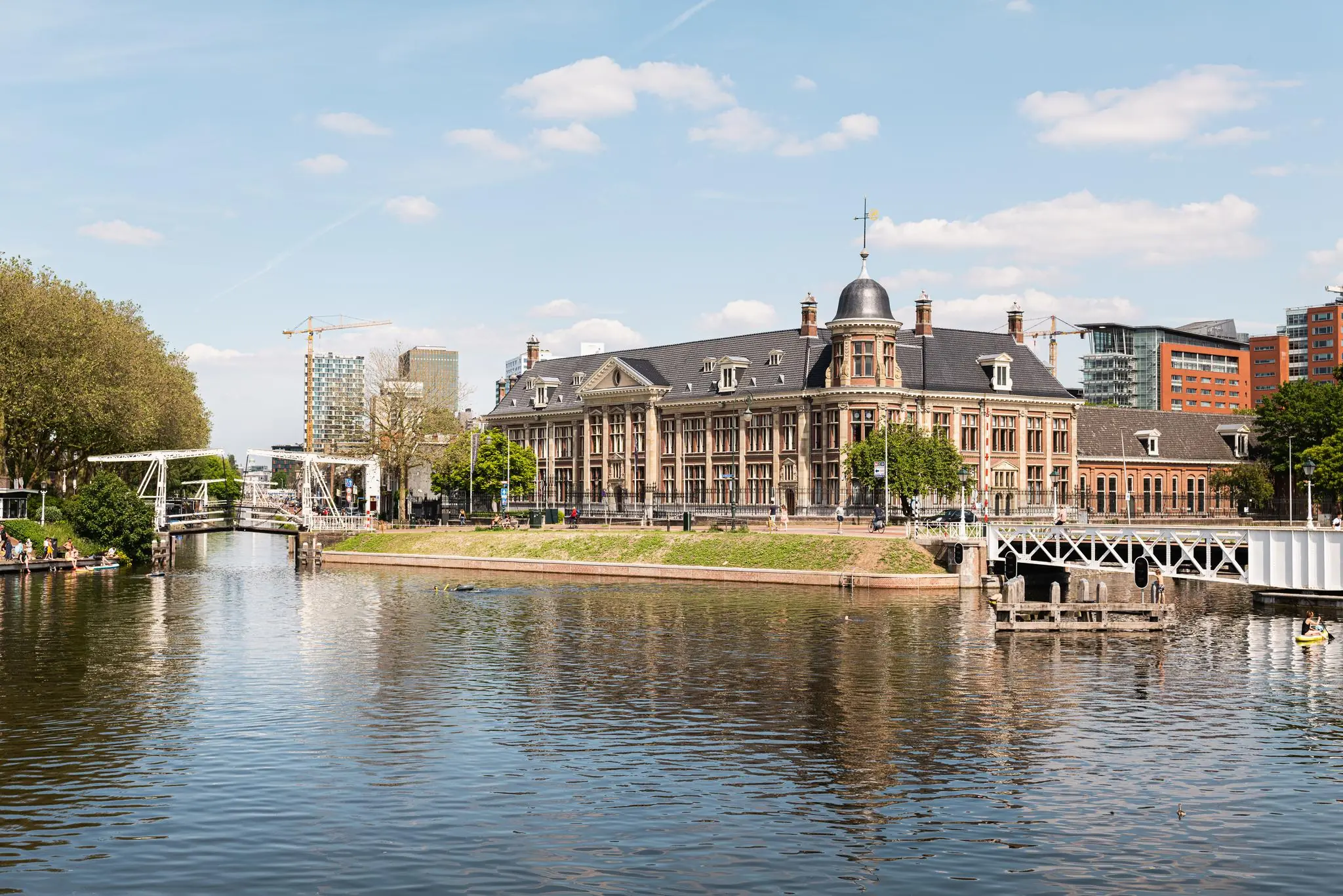 Historisch gebouw aan de Muntkade in Den Haag met bruggen en moderne hoogbouw op de achtergrond, gezien vanaf het water.