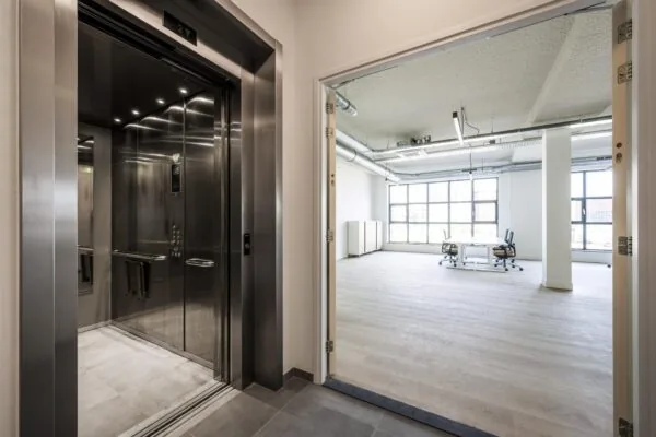 Een openlift op een moderne kantoorverdieping met zicht op een lichte, lege kantoorruimte met bureaustoelen en grote ramen.