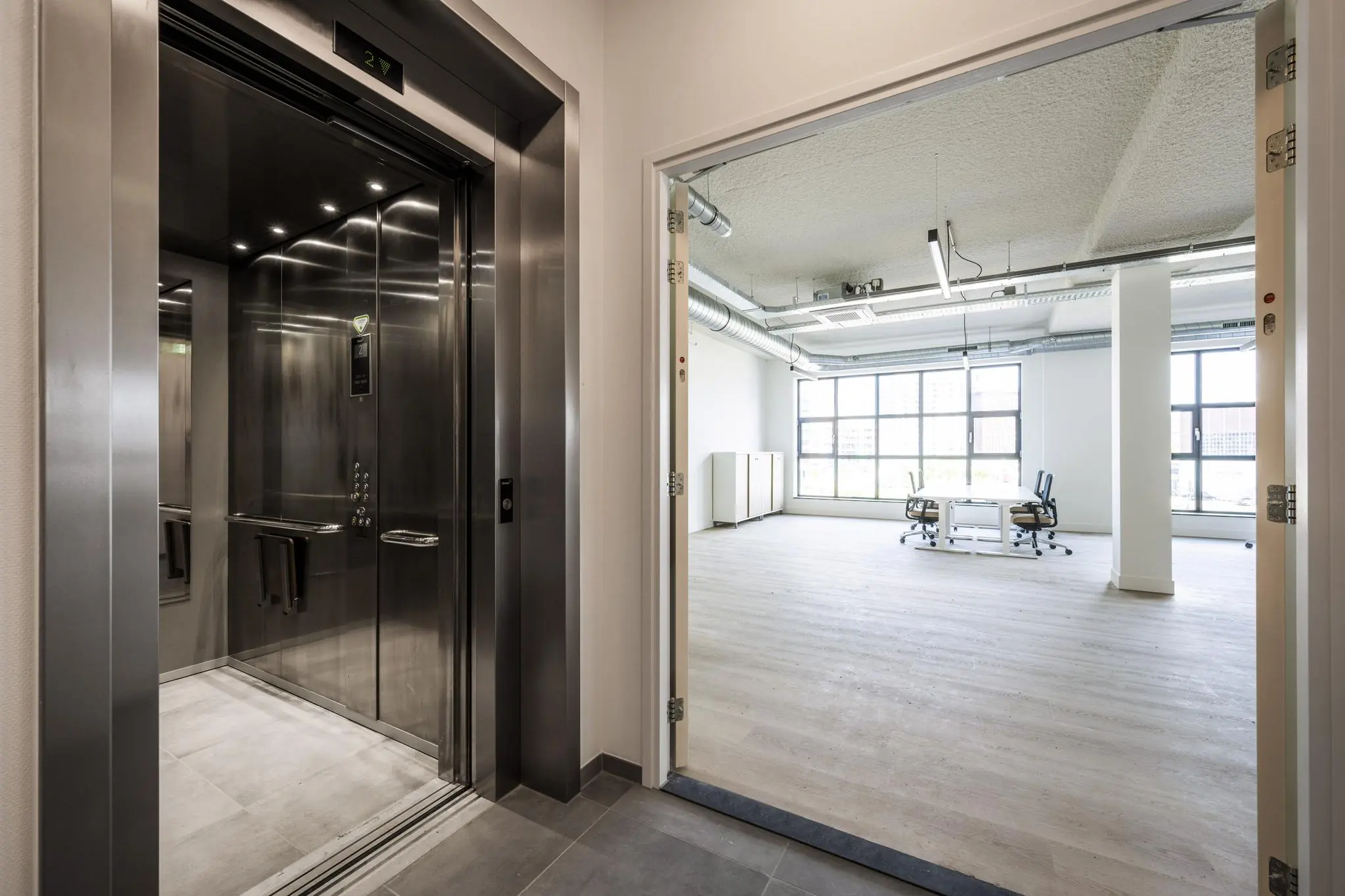 Een openlift op een moderne kantoorverdieping met zicht op een lichte, lege kantoorruimte met bureaustoelen en grote ramen.