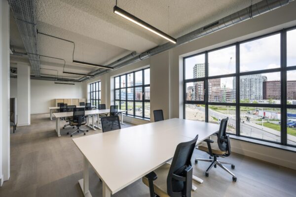 Moderne, lichte kantoorruimte met meerdere bureaus en grote ramen met uitzicht op nieuwbouw rondom de Hellingbaan.