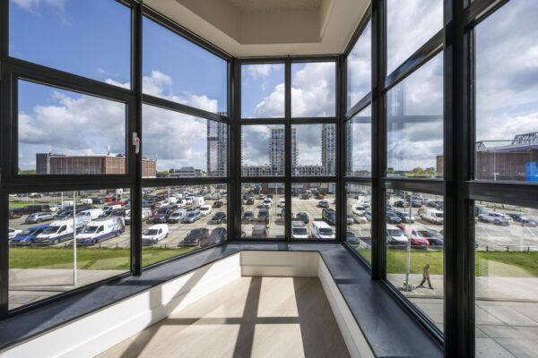 Uitzicht vanuit een glazen erker op een parkeerterrein en moderne hoogbouw bij de Hellingbaan.