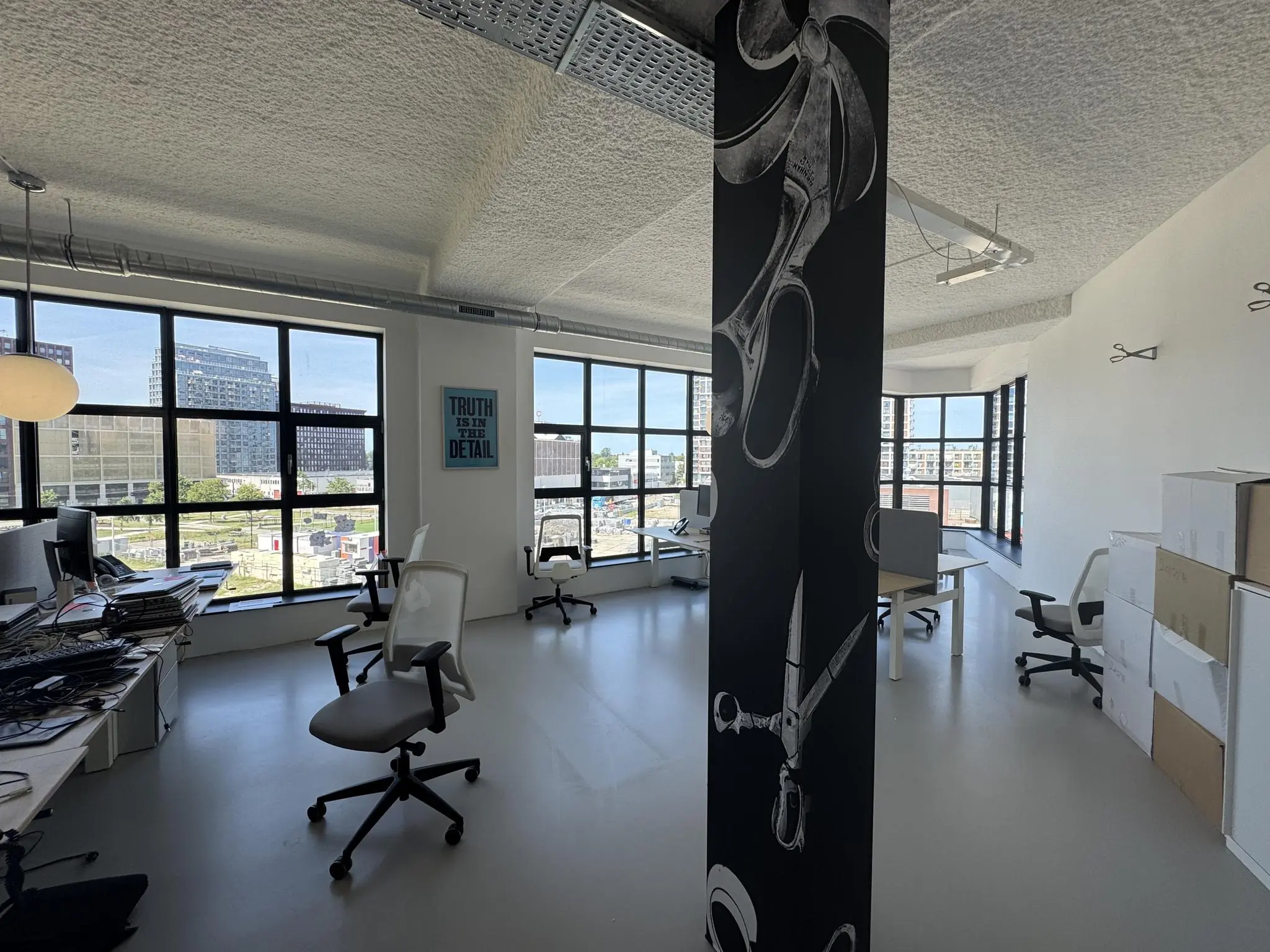 Modern en licht kantoor met bureaus, designstoelen en een zwarte zuil met schaarillustraties, met uitzicht op de stad via grote ramen.