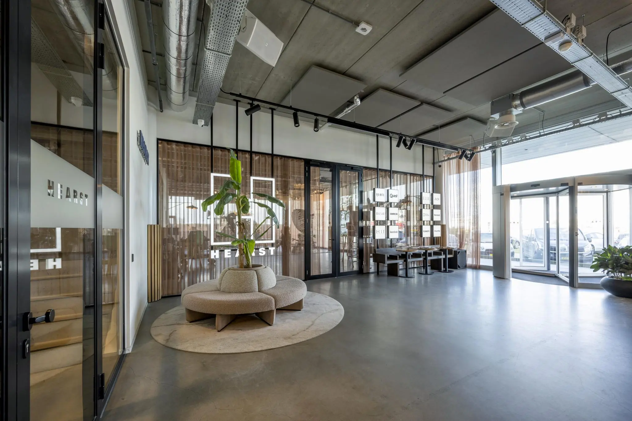 Moderne en lichte ontvangsthal van het Hearst-kantoor aan de Moermanskkade, met glazen wanden, industriële elementen en een ronde zitplek met grote plant.