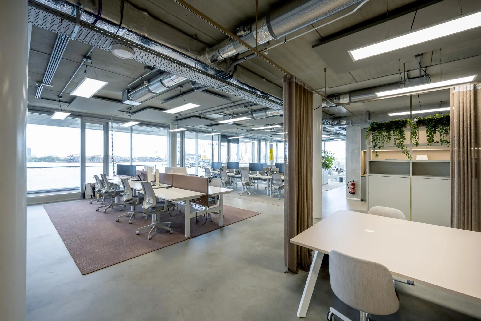 Modern open kantoorinterieur aan de Moermanskkade met werkplekken, industriële plafonds en uitzicht op het water.