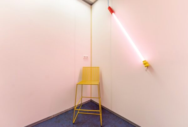 Geel metalen krukje in een hoek van een kamer met een diagonaal gemonteerde neonlamp aan de muur op Daltonlaan.