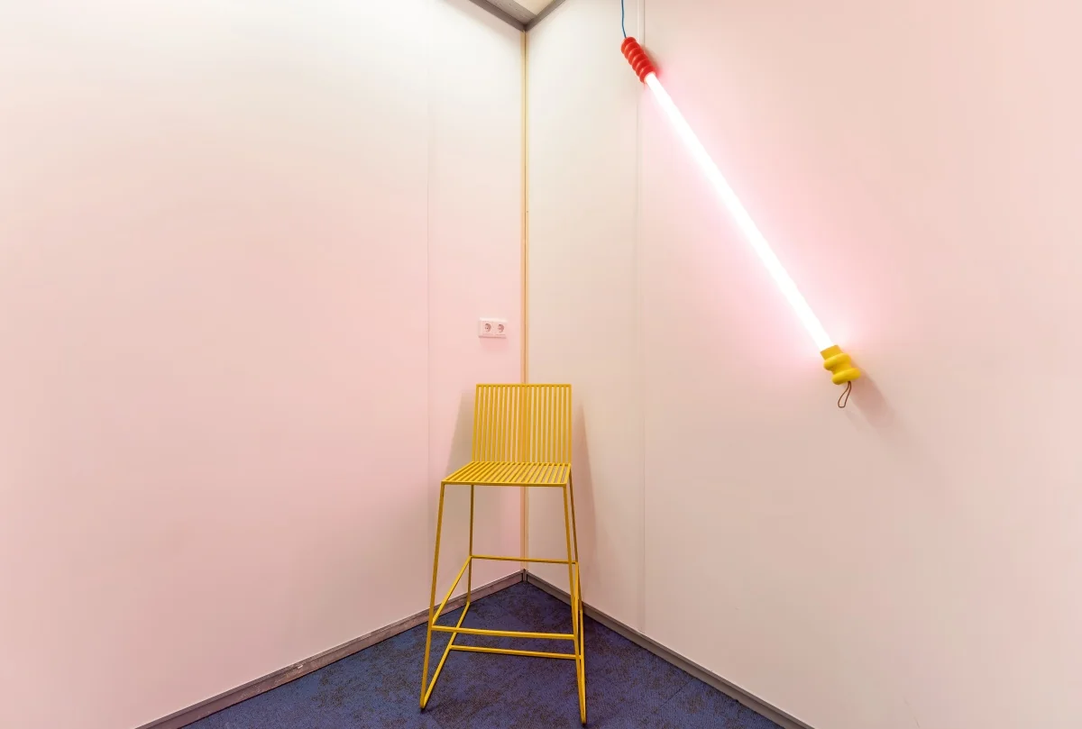 Geel metalen krukje in een hoek van een kamer met een diagonaal gemonteerde neonlamp aan de muur op Daltonlaan.