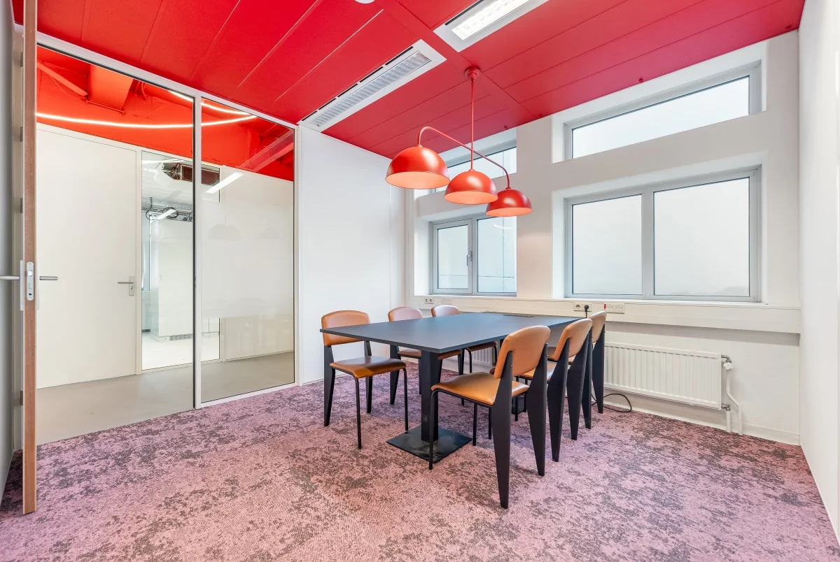 Vergaderruimte aan de Daltonlaan met rood plafond, moderne verlichting, zwarte tafel en zes stoelen op een paars tapijt.