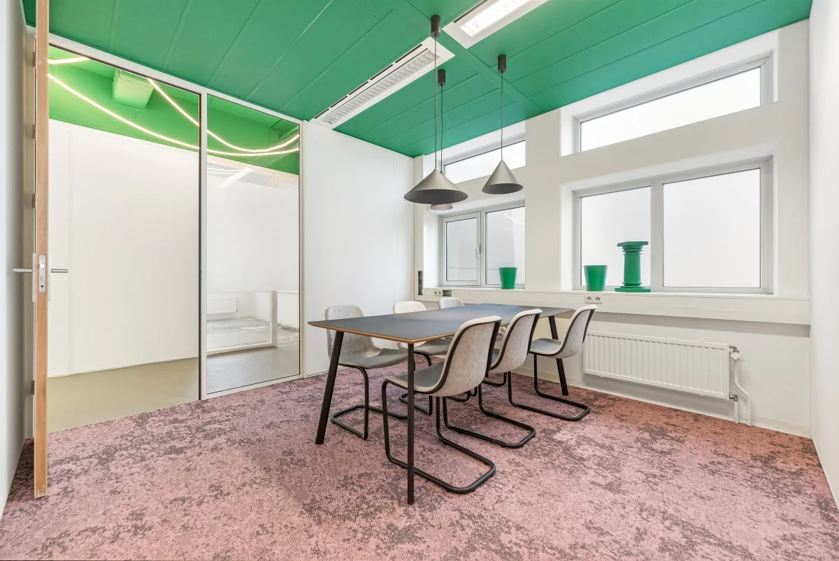 Moderne vergaderruimte aan de Daltonlaan met groen plafond, roze vloerbedekking en een strakke inrichting met zes stoelen en hanglampen.