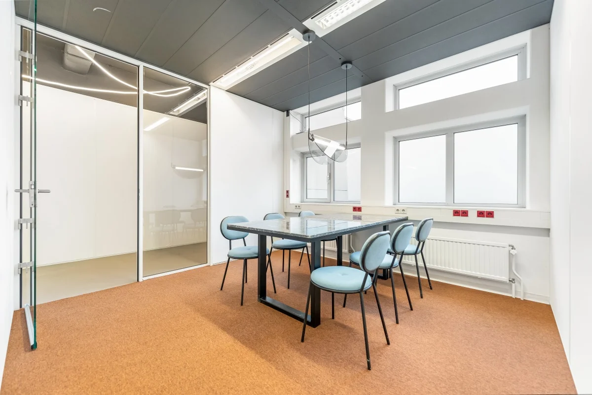Vergaderruimte aan de Daltonlaan met glazen wanden, moderne verlichting, en een tafel met zes lichtblauwe stoelen.