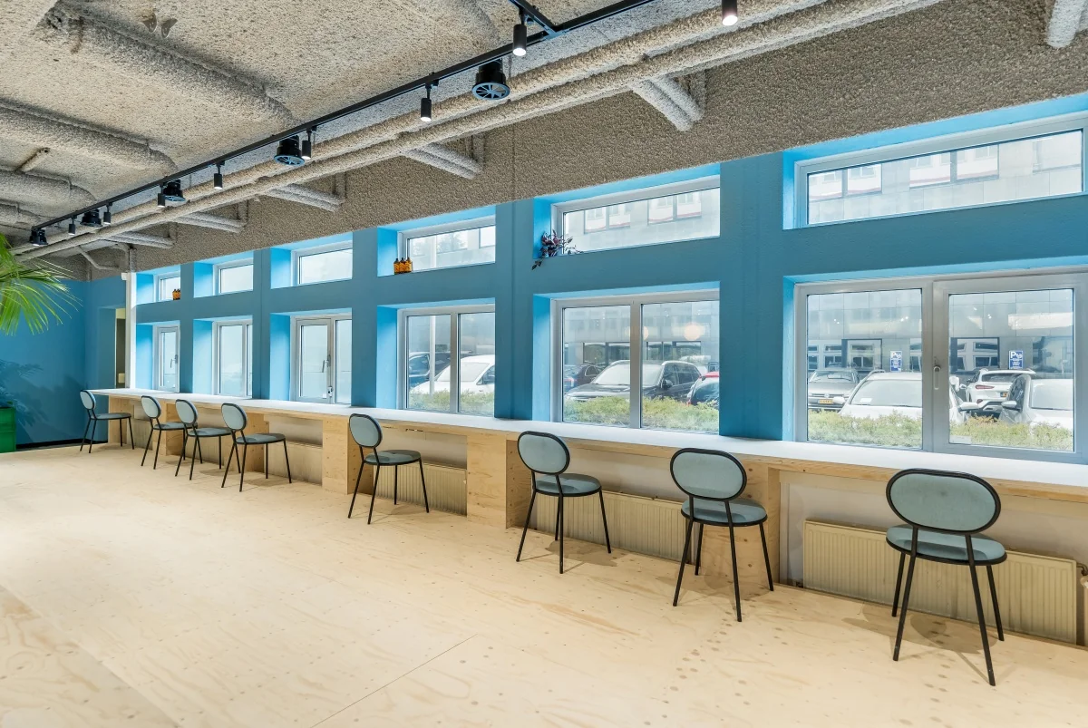 Moderne werkruimte aan de Daltonlaan met houten vloer, lange werktafel, blauwe stoelen en grote ramen met uitzicht op geparkeerde auto's.
