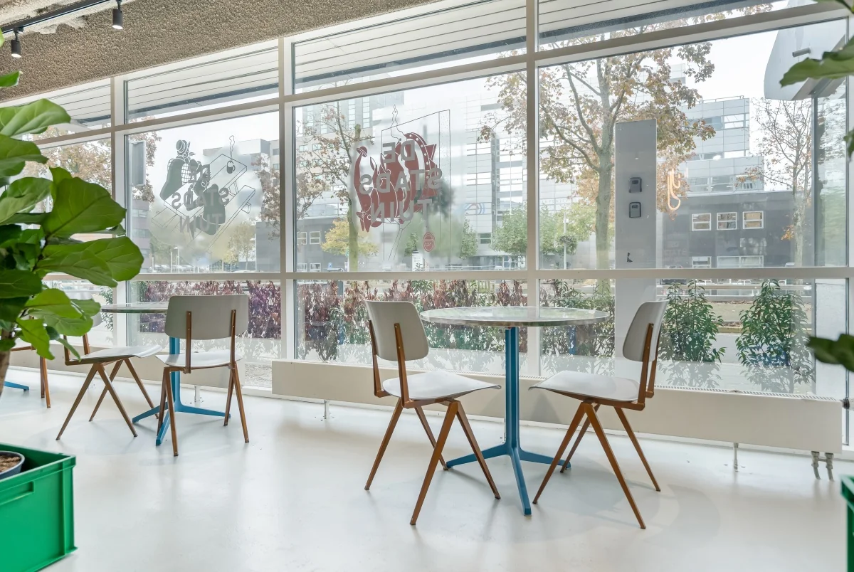 Moderne en lichte zithoek met ronde tafels en houten stoelen aan de Daltonlaan, met uitzicht op kantoorgebouwen.