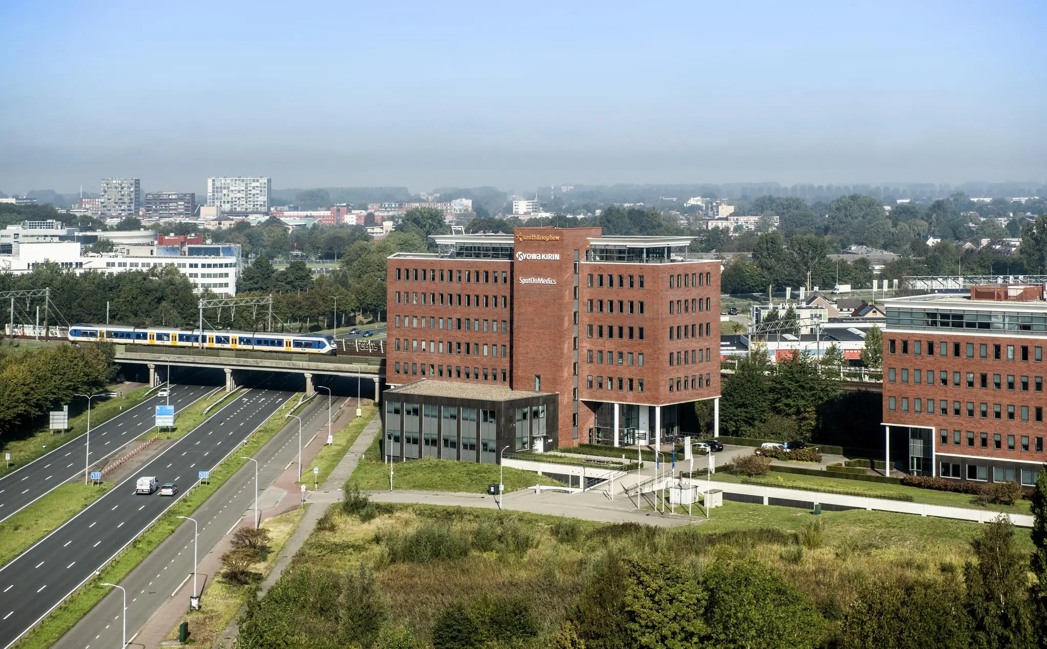 Zicht op een rood bakstenen kantoorgebouw met de namen Smith+Nephew, Kyowa Kirin en SpotOnMedics langs een snelweg en spoorlijn met passerende trein.