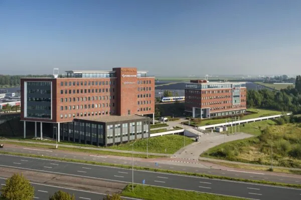 Luchtfoto van een modern kantorencomplex met rode bakstenen gebouwen langs een snelweg en een passerende trein op de achtergrond.