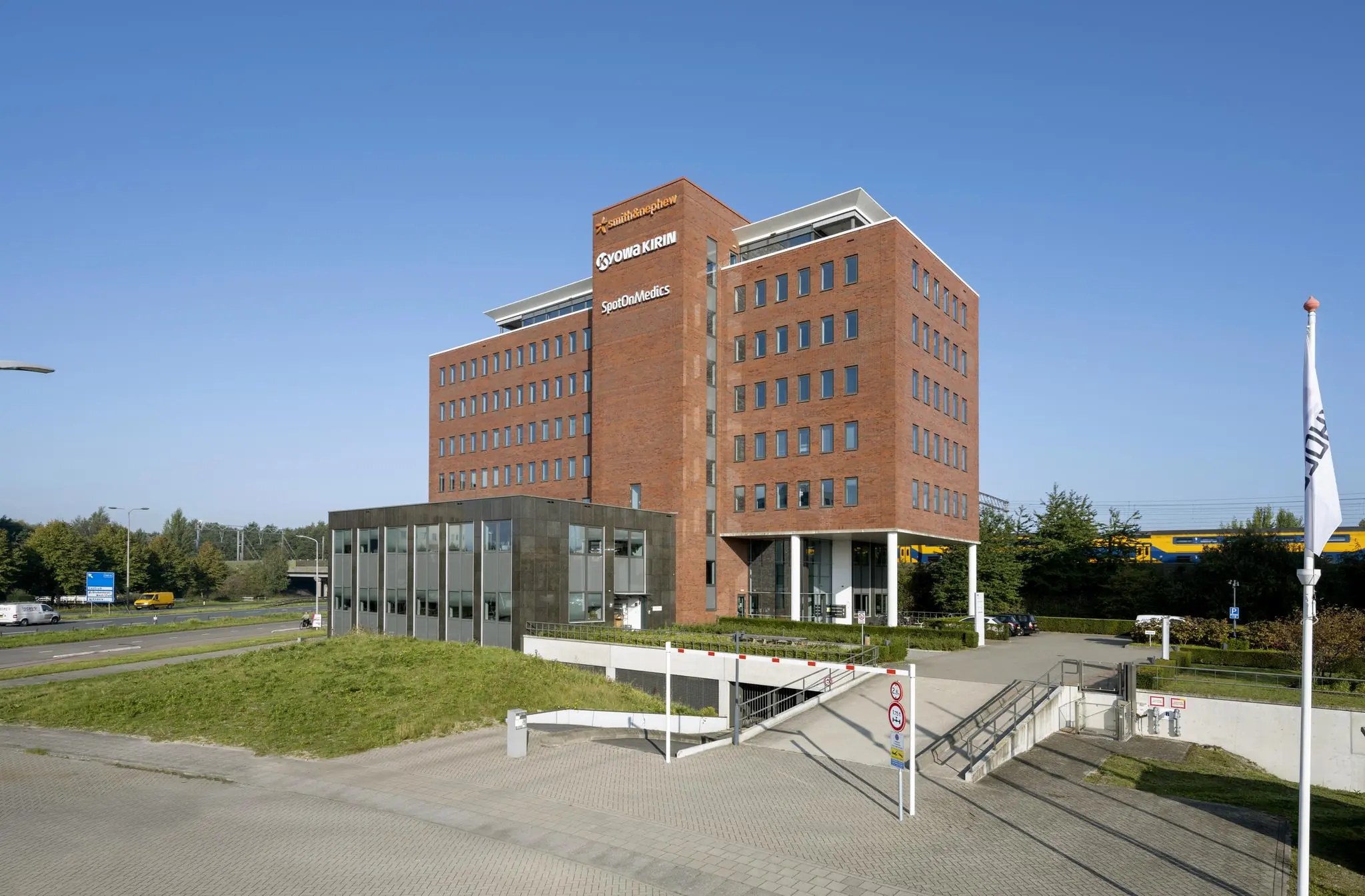 Kantoorgebouw met de namen Smith+Nephew, Kyowa Kirin en SpotOnMedics langs een spoorlijn op een zonnige dag.