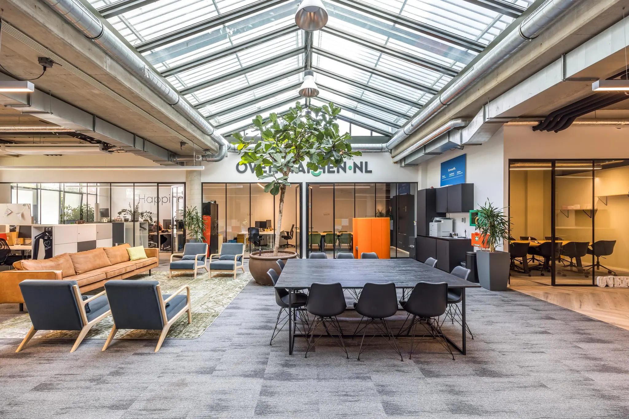 Modern en licht kantoorinterieur met zithoek, vergadertafels, veel glas en een grote plant onder een glazen dak bij Overtoom.nl.