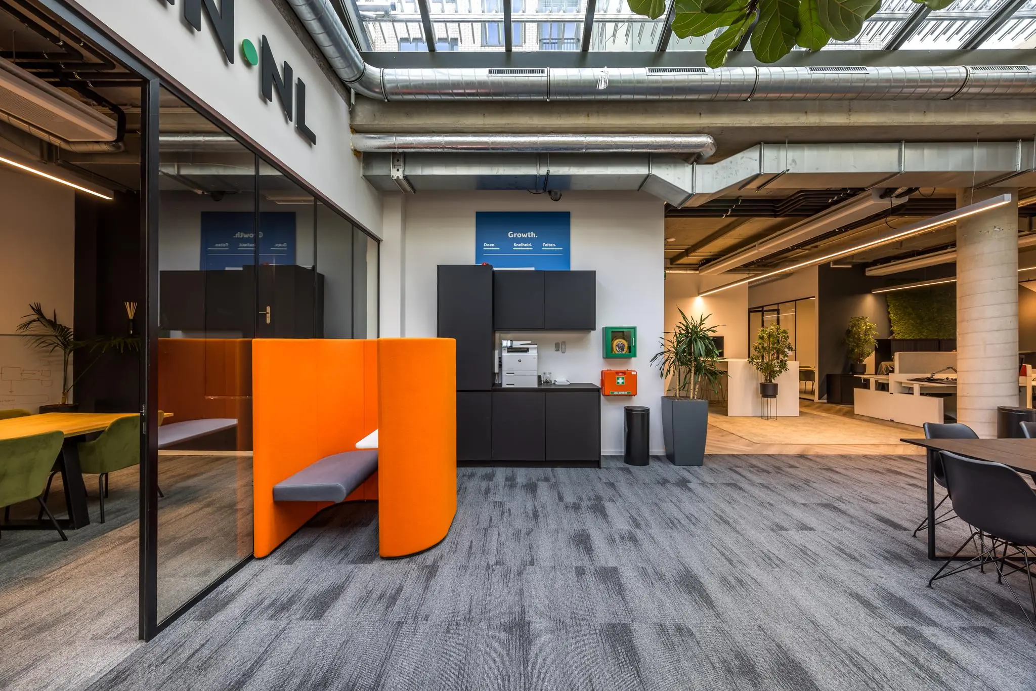 Moderne kantoorruimte aan de Overtoom met een opvallende oranje zithoek, planten, en een koffiecorner onder een glazen dak.