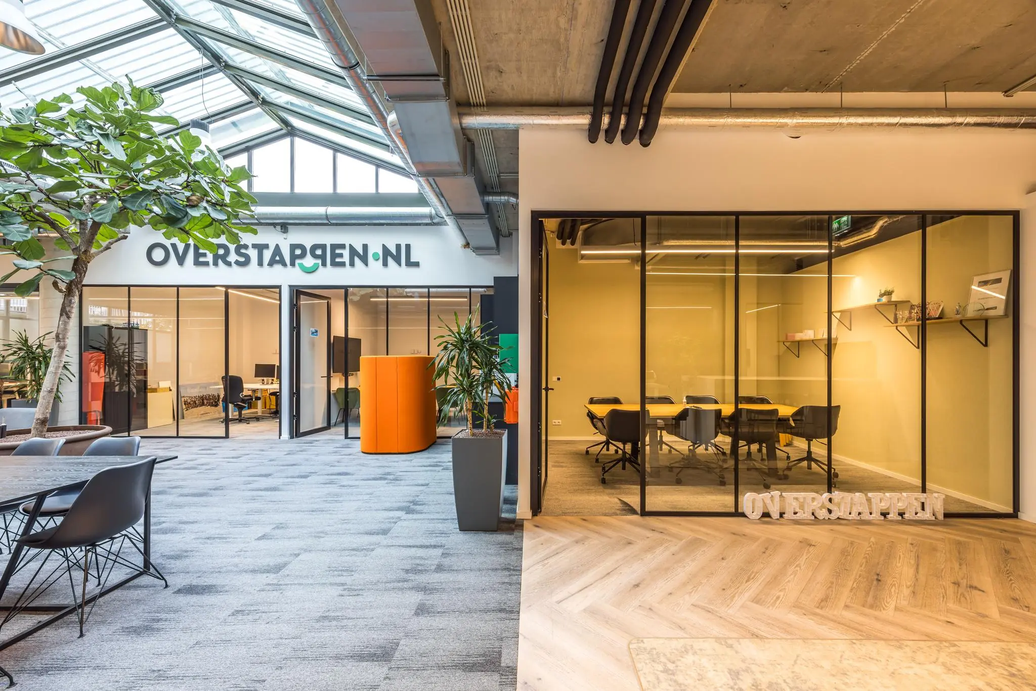 Moderne kantoorruimte van Overstappen.nl met glazen vergaderruimtes, planten en een grote lichtinval via het glazen dak.