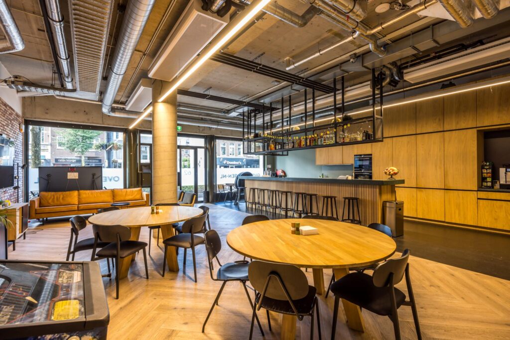 Moderne lounge- en eetruimte met houten tafels, een bar en zicht op de Overtoom in Amsterdam.