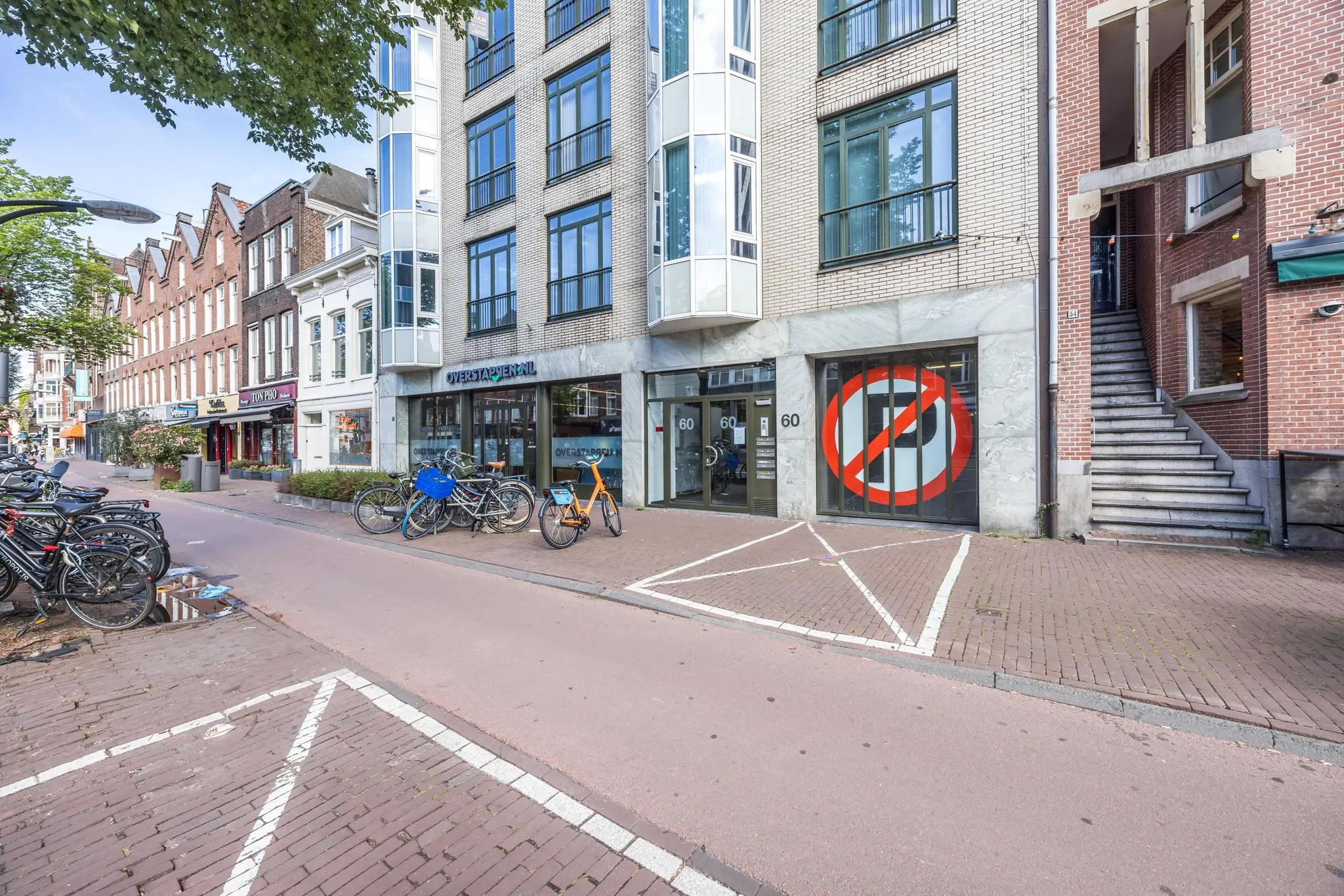 Straatbeeld van de Overtoom in Amsterdam met winkels, geparkeerde fietsen en het pand op nummer 60 met opvallend verkeersbord op de gevel.