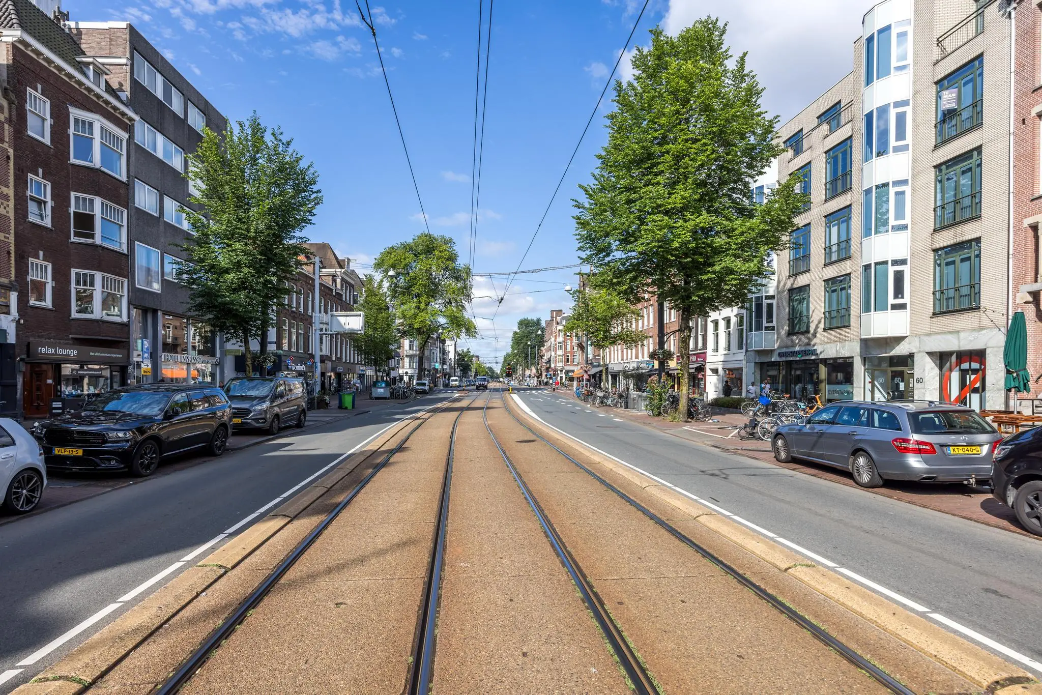 Straatbeeld van de Overtoom in Amsterdam met trambaan, geparkeerde auto's en winkels aan weerszijden.