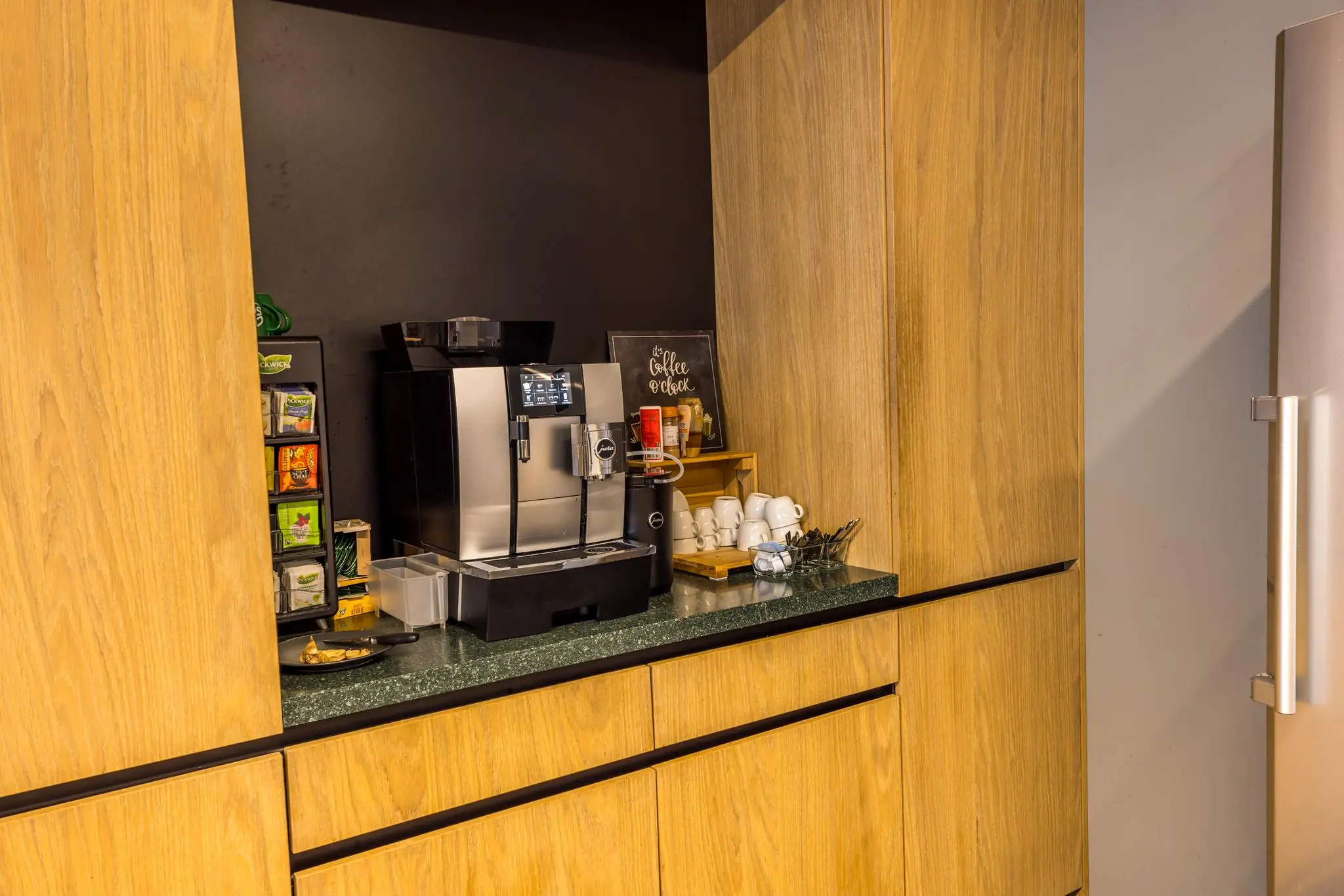 Moderne koffiehoek met een volautomatische koffiemachine, theeselectie en servies in een houten keukenmeubel.