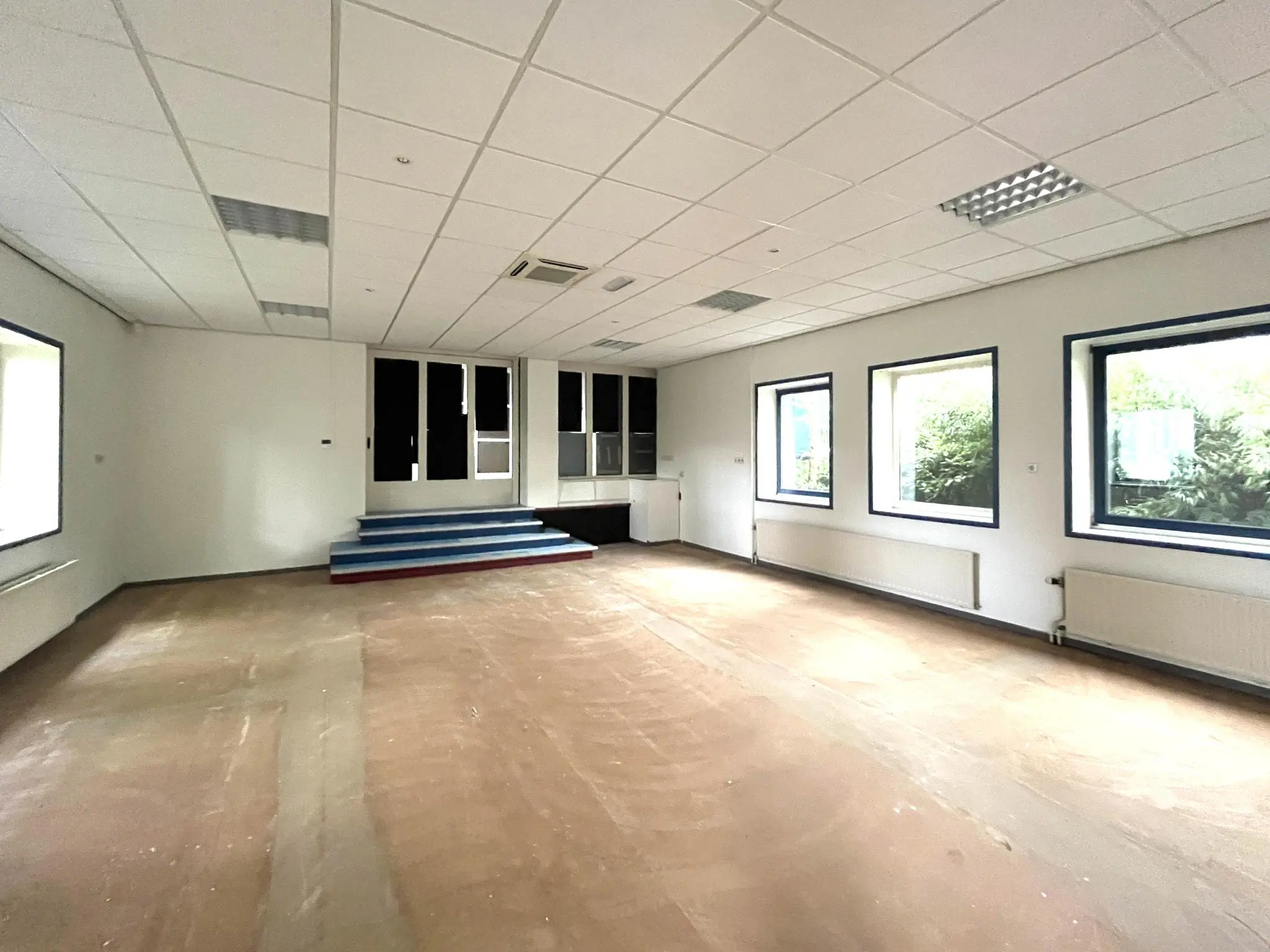 Leegstaand kantoor of klaslokaal aan de 2e Daalsedijk met witte wanden, systeemplafond, grote ramen en een klein verhoogd podium.