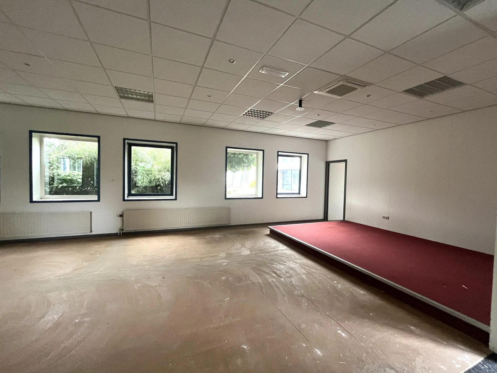 Leegstaand kantoorruimte aan de 2e Daalsedijk met drie ramen en een verhoogd podium met rode vloerbedekking.