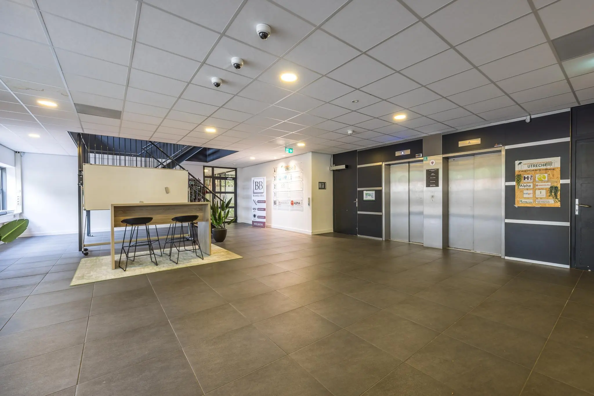 Moderne en ruime entreehal van een kantoorgebouw aan de Daltonlaan met liften, bewegwijzering en een zit-werkplek.