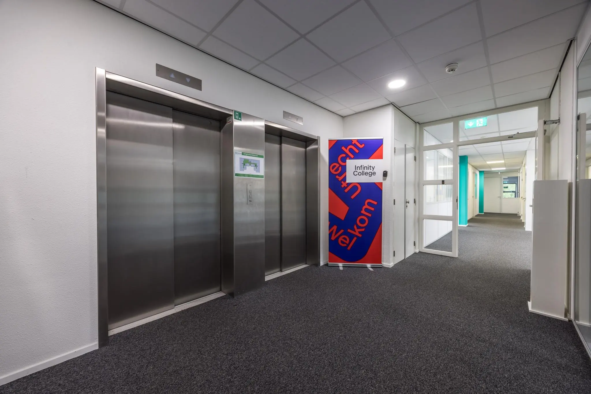 Hal met twee roestvrijstalen liften en een welkomstbord van Infinity College in het gebouw aan de Daltonlaan.